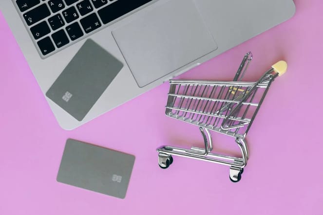 Imagem de um fundo rosa com carrinho de compras em miniatura ao lado de um notebook e dois cartões de crédito