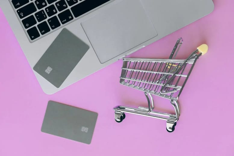 Imagem de um fundo rosa com carrinho de compras em miniatura ao lado de um notebook e dois cartões de crédito